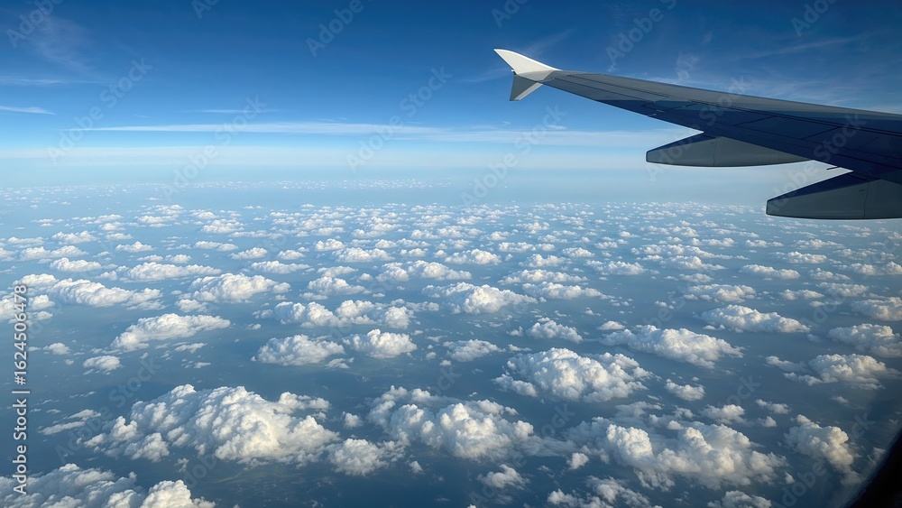 Fototapeta premium Quiet flight window outlook showing plane wing, billowy clouds beneath, and clear azure sky above, stretching over a vast patterned scenery.