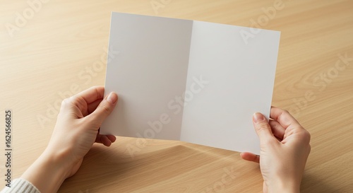 A blank white card is held open by two hands against a light wood background