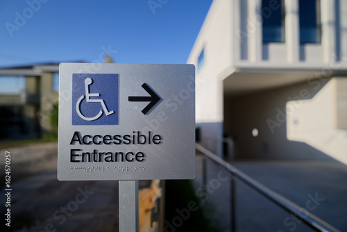 Rectangular accessible entrance sign, featuring blue wheelchair disability icon, black text, braille, and black arrow outside the entry to a building