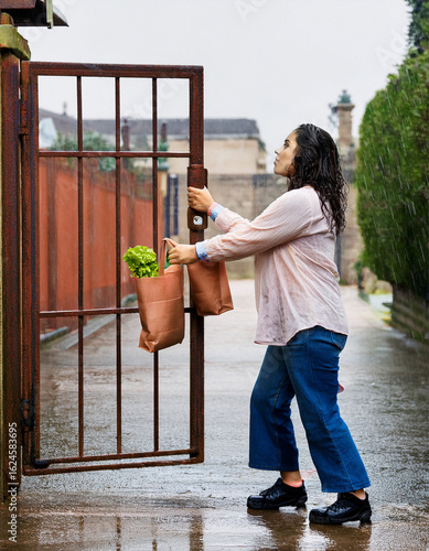 A young woman in her late 20s struggling to open a rusty metal gate in the rain. She's holding a grocery bag in one hand, hair wet, her other hand gripping the old iron latch tightly. 