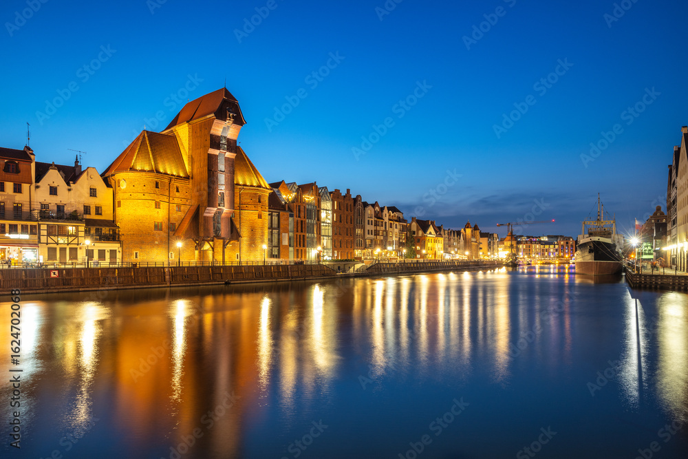 Naklejka premium Gdansk with medieval port crane over The Motlawa river, a city on the Baltic coast of northern Poland, Europe. Night Cityscape: Reflections of old buildings on a river under the dark blue sky.