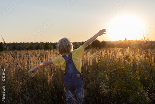 child joyfully looks at the setting sun, arms outstretched standing on a meadow and bathing in the sun's rays. Happy childhood. Forward to a bright future. Back view. Backlight and soft focus.