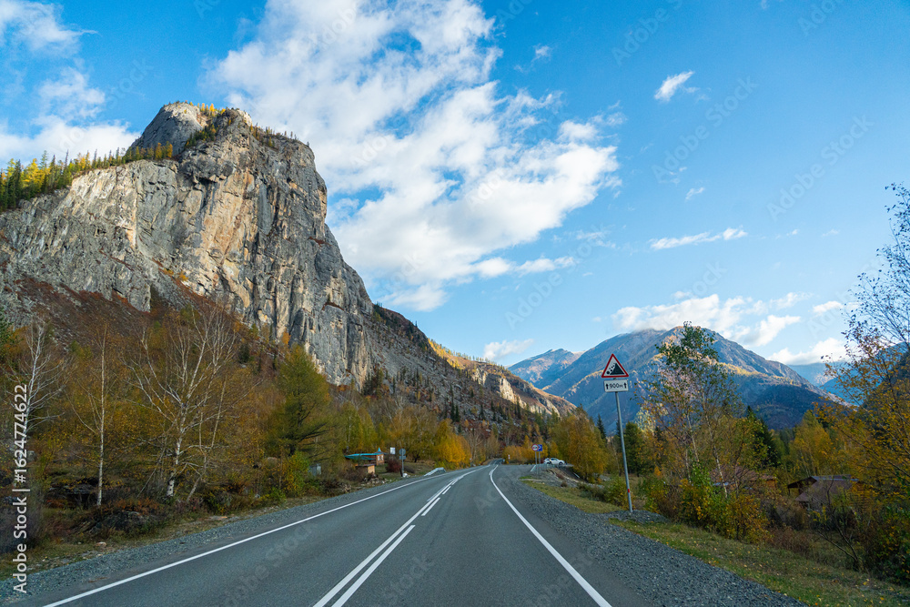 Fototapeta premium A winding road leads into the distance with a tall, rocky mountain on the left side and a forest of autumn trees on the right