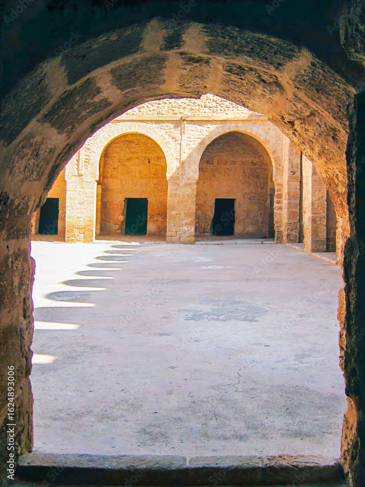 Naklejka premium Sunlight cuts through arched silence — a glimpse into the ribat’s inner courtyard in Sousse. Light and shadow mark sacred geometry in ancient stone.