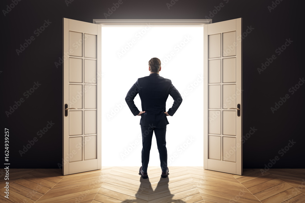 Businessman standing with hands on hips in dark room facing open double doors with bright light, symbolizing opportunity and future success. Businessman standing with hands on hips in dark room facing open double doors with bright light, symbolizing opportunity and future success.