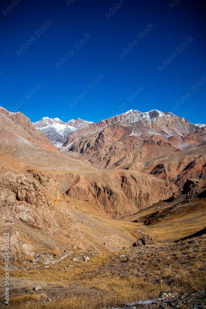 Fototapeta premium A vertical view of a vast, brown and golden mountain valley with rugged mountains in the distance and a clear blue sky