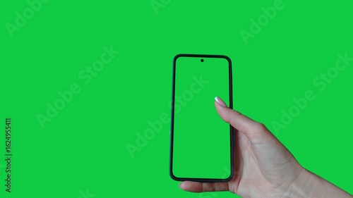 Young woman hand touch smartphone with green screen on green background. Phone in a female hand. Scrolling and swiping on a green phone screen. Template for scrolling through a feed in social network