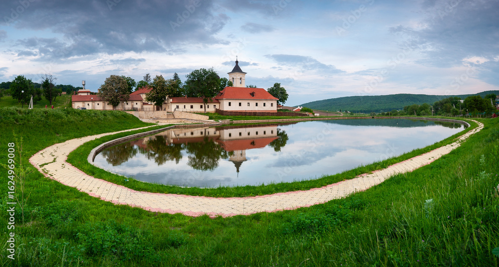 Obraz premium A tranquil panoramic view of a monastery with red-tiled roofs and a bell tower, beautifully reflected in a still pond, representing religious architecture and serene landscapes for travel and spiritua