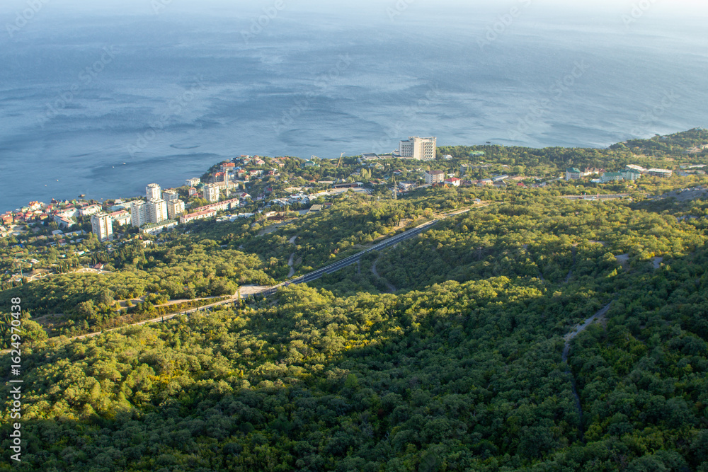 Obraz premium An aerial view of a coastal city nestled among lush green trees, with modern buildings and a winding road overlooking a calm sea, perfect for depicting a travel destination, urban planning, or nature