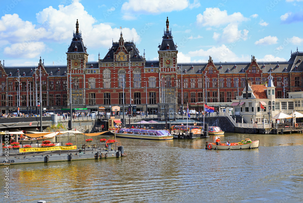 Obraz premium Amsterdam Central Station located very close to the River Amstel in the City Centre. There are many tourist boats eager to take tourists on a trip around the canals.