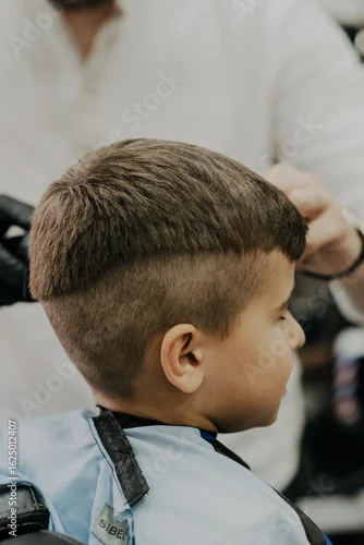 Fototapeta samoprzylepna Smiling little boy sitting at barber shop, while hairdresser styling his hair.