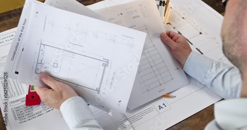 Experienced man architect sits at desk and examines sketches of different apartment rooms. Office worker in shirt pays close attention to details in bedroom layout
