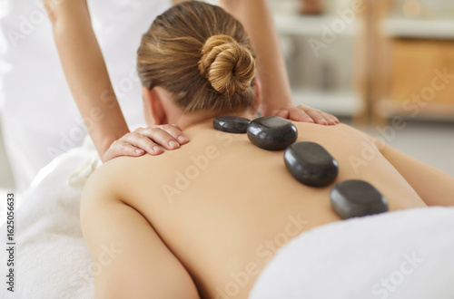 Beautiful young woman lying on massage table and receiving relaxing back hot stone massage therapy in spa center. Relaxed girl enjoying wellness therapy from professional masseur in beauty clinic.