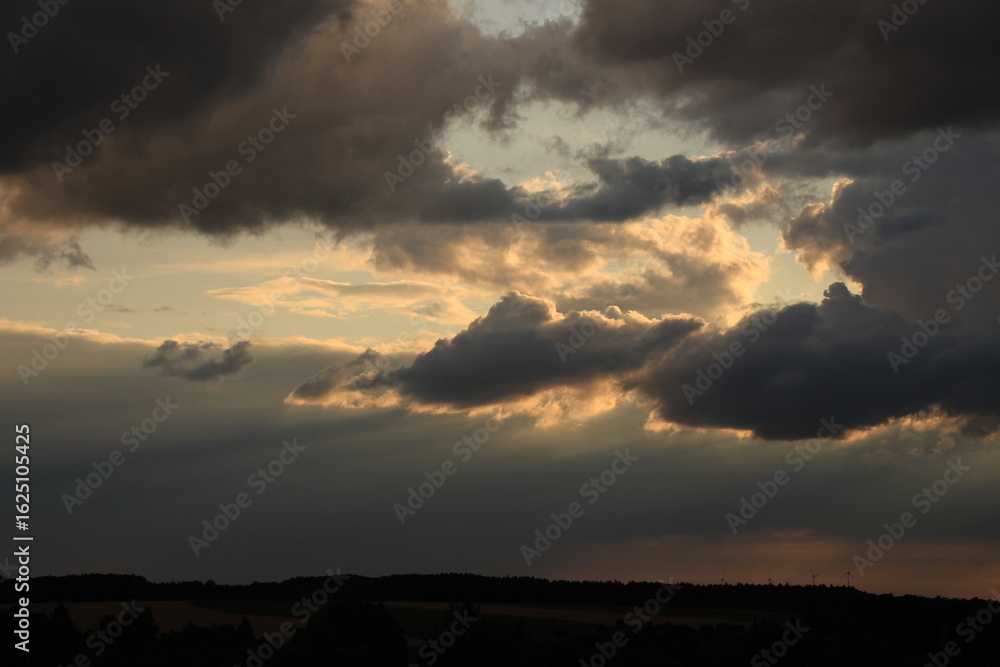 Atmospheric landscape featuring a dramatic sunset sky filled with dark storm clouds and glowing rays of evening light breaking through. The silhouette of rolling hills and distant wind turbines adds a Atmospheric landscape featuring a dramatic sunset sky filled with dark storm clouds and glowing rays of evening light breaking through. The silhouette of rolling hills and distant wind turbines adds a