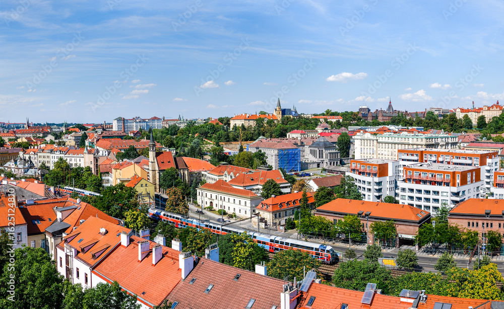 Obraz premium Panoramic view of Prague cityscape with red-tiled rooftops, residential buildings, and a commuter train passing through the urban area on a sunny summer day.