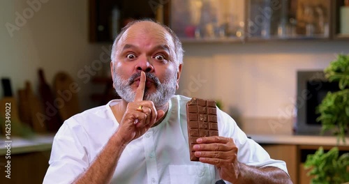Senior Indian man secretly eating chocolate brick in modern kitchen, Asian elderly foodie sneaking thick chocolate slab with mischievous expression, enjoying sweet treat alone at home