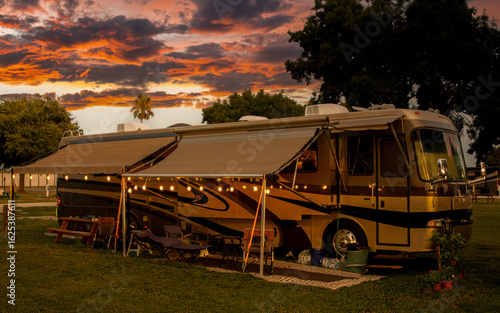 A motorhome is parked on a grassy field, with a canopy extending out, under which there are string lights and seating. The sky above is filled with dramatic, colorful clouds typical of a sunset or sun