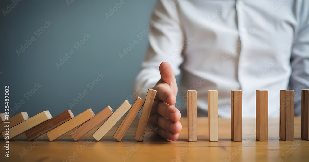 © fatemaakter - Preventing Domino Effect: A person's hand halting the cascading collapse of a row of wooden blocks, symbolizing risk management and strategic intervention. © fatemaakter - Preventing Domino Effect: A person's hand halting the cascading collapse of a row of wooden blocks, symbolizing risk management and strategic intervention.