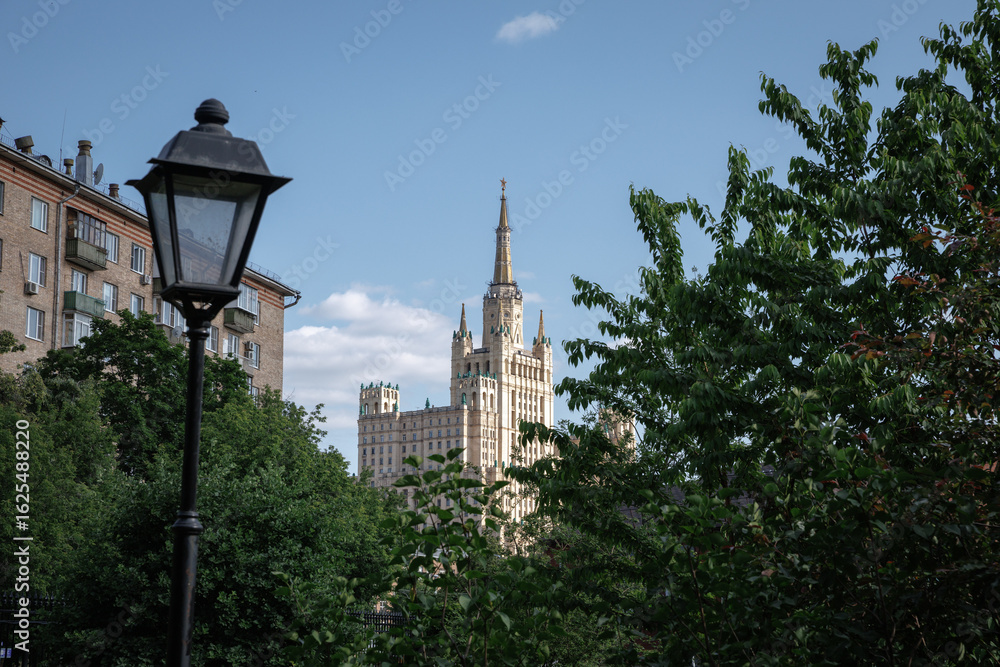 Fototapeta premium Urban scene featuring a tall building behind green foliage and a streetlight under a clear blue sky. The architecture and greenery create a harmonious blend of urban and natural elements.