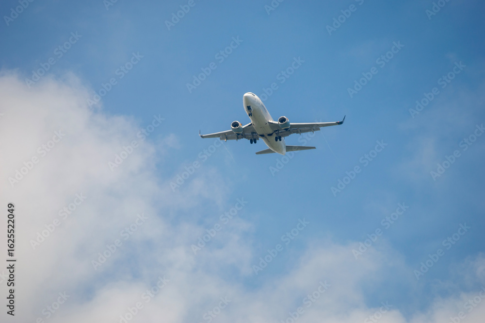 Obraz premium Avión volando en un cielo azul con nubes blancas mientras se aproxima a Puerto Rico. La imagen captura el momento de llegada a la isla caribeña, simbolizando viajes, turismo y transporte aéreo.