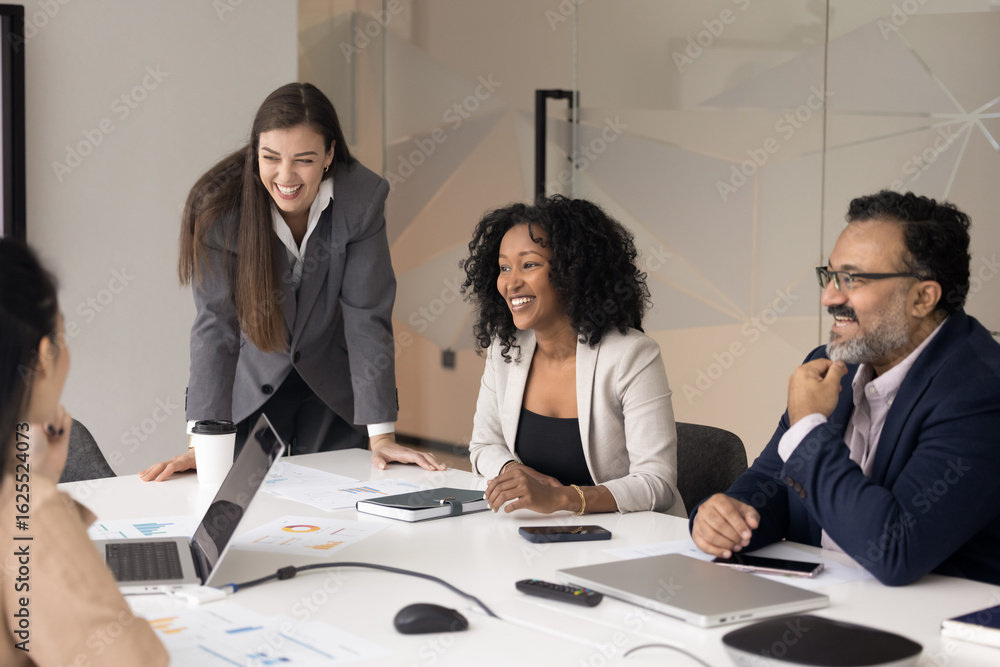 Fototapeta premium Joyful collaboration. Smiling young woman ceo boss and multiracial team sales business development professionals engage in laughter on boardroom meeting listen to coworker giving laptop presentation