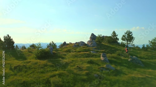 Runner in vibrant gear runs through a lush, mountainous trail, showcasing endurance. Verdant landscape with rocky outcrops and pine trees under a clear sky.