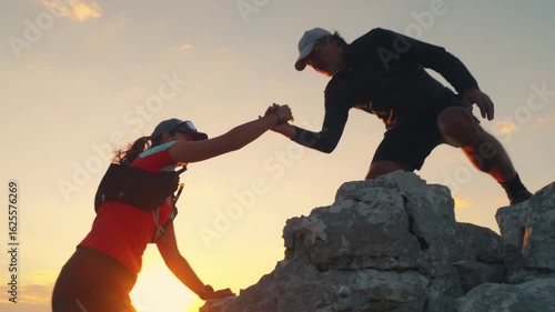 Two runners ascend a rocky peak at sunset, one helping the other. Their determined expressions and teamwork highlight endurance and achievement in a stunning natural setting.