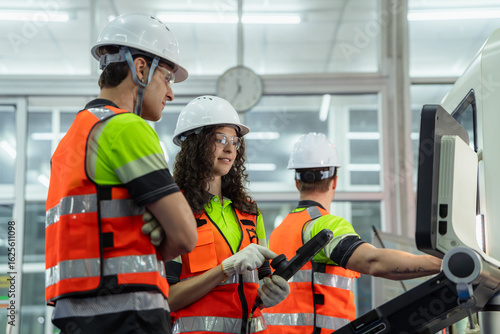 Three people wearing safety gear and orange vests. Group of machinists in an on-the-job training session, learning to program and control an automated industrial machine.