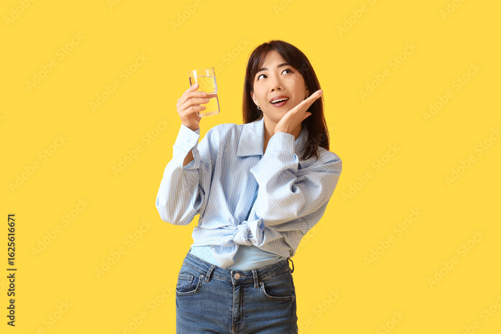 Happy young Asian woman with glass of water on yellow background Happy young Asian woman with glass of water on yellow background
