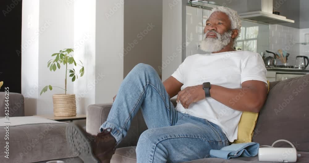 Senior African American man feeling stiff shifting on couch grabbing glass and scrolling smartphone Senior African American man feeling stiff shifting on couch grabbing glass and scrolling smartphone