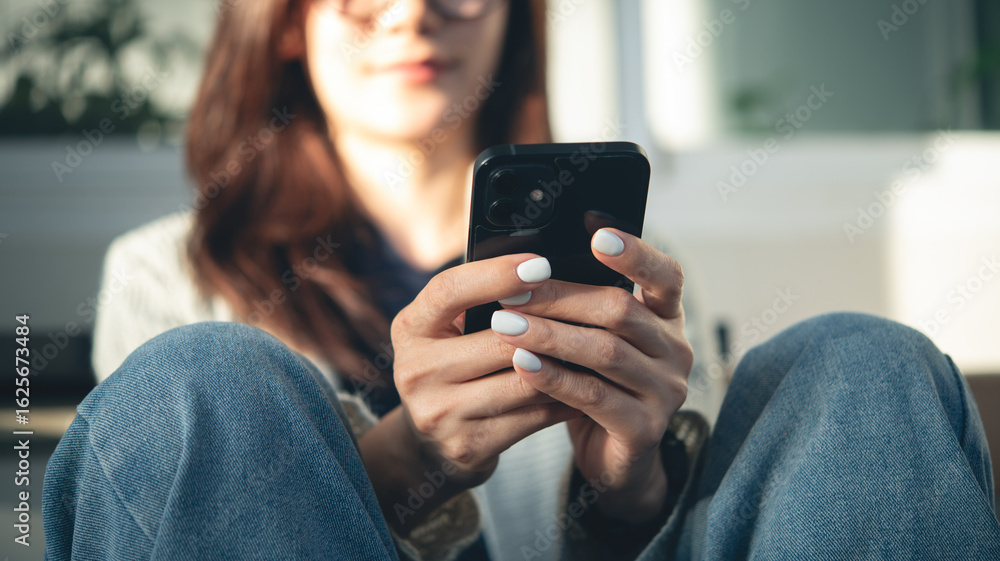 Fototapeta premium Close-up, young hipster girl sitting on the floor at home interior, using smartphone for social network or online shopping via mobile app. Young asian woman typing text message via mobile phone