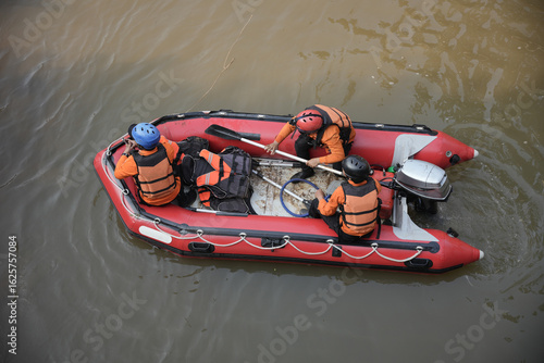 Rescuers in orange uniforms and helmets row a red rubber boat in murky water during a search and rescue operation. Concept photo of flooding and river tracing.