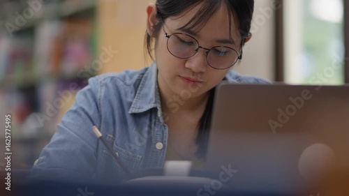 Young Asian woman, early 20s, wearing glasses and casual clothes, studying in a library using a laptop and book. Ideal for concepts like education, learning, focus, university, or academic life.