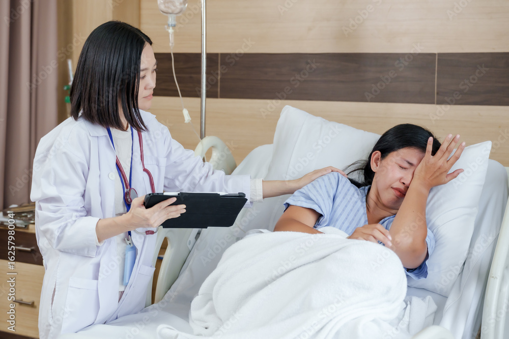 Fototapeta premium Female patient on hospital bed showing distress as asian female doctor gently comforts and listens while holding tablet during emotional consultation