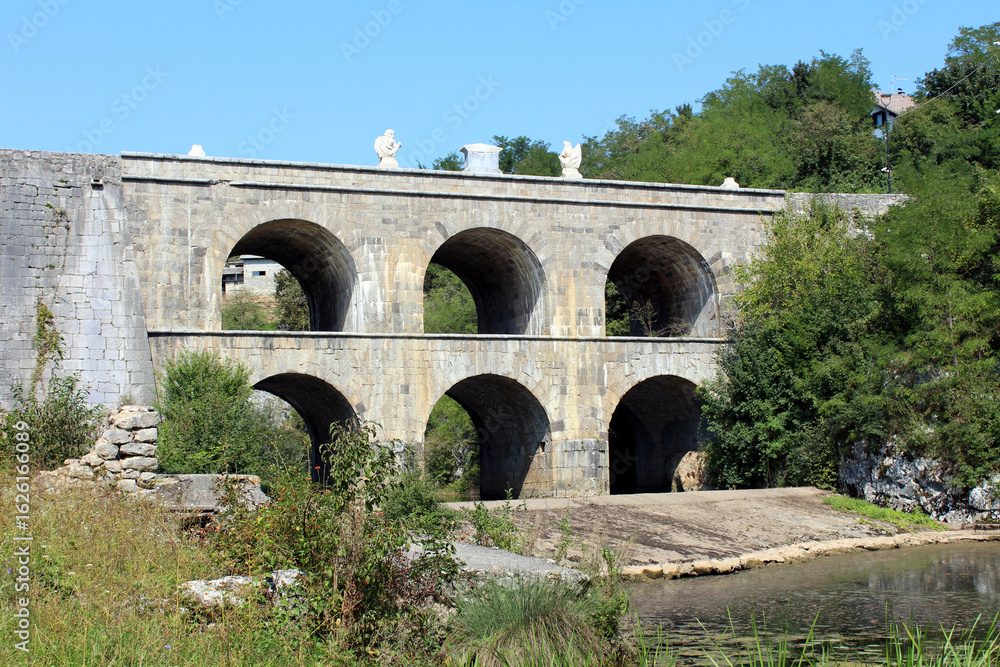 Fototapeta premium A multi-level stone aqueduct, featuring two rows of large, symmetrical arches, stands over a river, showcasing impressive historic masonry in a green, rural landscape