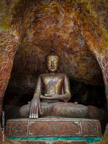 A Buddha statue in a meditation pose, seated inside a natural cave. The sculpture shows the Earth-touching gesture, symbolizing enlightenment, surrounded by rich textures and colors.