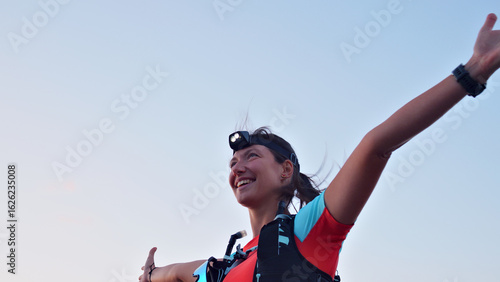 Joyful Caucasian female runner celebrating achievement during cross-country run, wearing headlamp and hydration pack, arms raised against clear sky, embodying endurance and sport spirit.