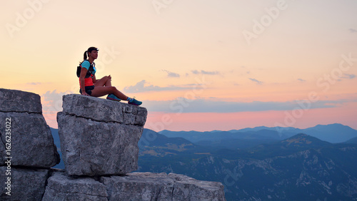 Canvas Print Determined female runner in vibrant gear atop a rugged cliff, overlooking a stunning mountainous landscape at sunset, embodying endurance and the joy of cross-country running