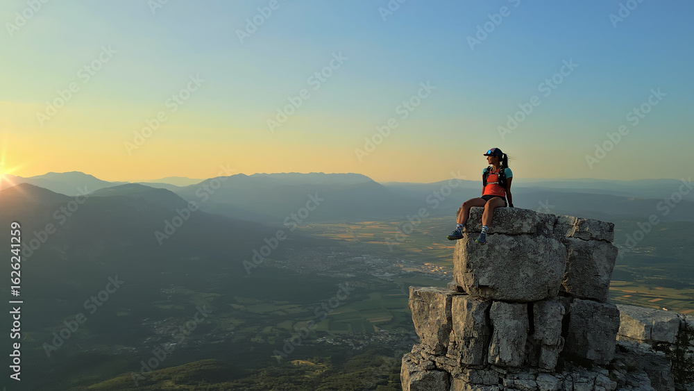 Fototapeta premium Hiker triumphantly sits on a rocky peak at sunrise, overlooking a vast valley. Environmental portrait captures achievement, endurance, and freedom, with golden light symbolizing perseverance.