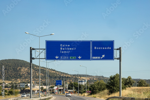 Highway signs showing directions to Ezine, Balıkesir, İzmir and Bozcaada, with countryside view under clear blue sky, Turkey