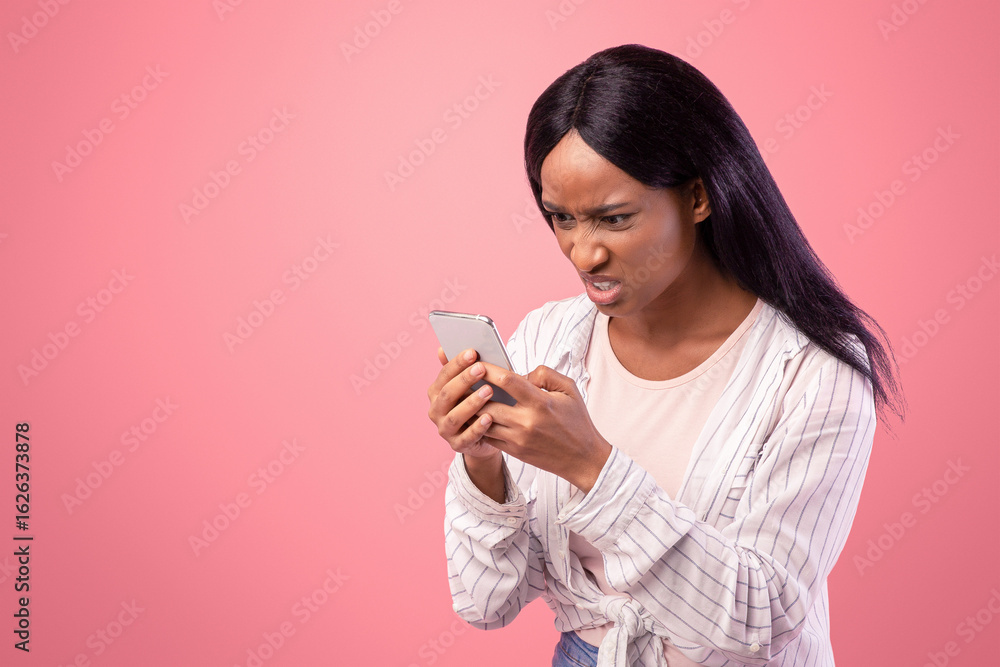 Angry black woman looking at her smartphone with disgust, reading annoying message or news on pink studio background. African American lady feeling stressed at social media publication Angry black woman looking at her smartphone with disgust, reading annoying message or news on pink studio background. African American lady feeling stressed at social media publication