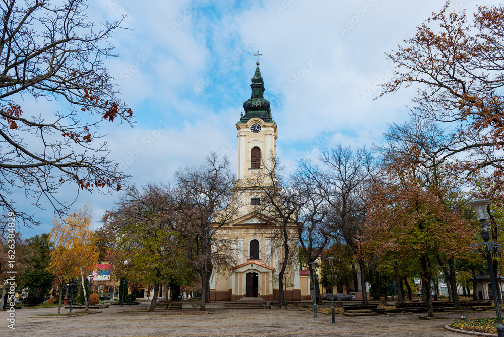 Obraz premium The Monastery of the Holy Trinity in Kikinda rises behind leafless autumn trees. Its baroque bell tower and quiet square reflect centuries of Serbian Orthodox tradition.