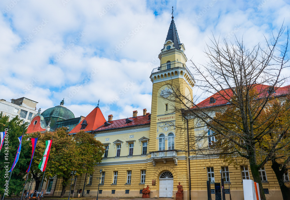 Obraz premium Kikinda City Hall stands as a historic landmark with a tall clocktower and red-tiled roofs. Its yellow facade and ornate details reflect classic Serbian civic architecture.