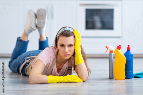Картината върху платно Tired young lady lying on floor among cleaning supplies, exhausted from housework in kitchen, copy space