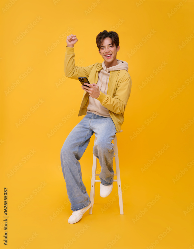 Fototapeta premium Handsome young happy smiling asian man celebrating online prize win using mobile phone sitting on chair isolated on yellow background.