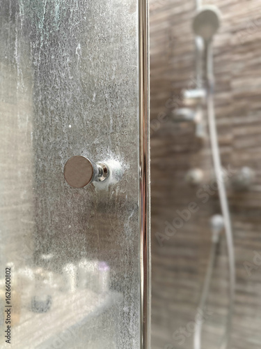Close-up of a dirty shower glass door with white mineral residue from hard water and soap scum. Blurred shower cabin in the background. Common bathroom cleaning issue.