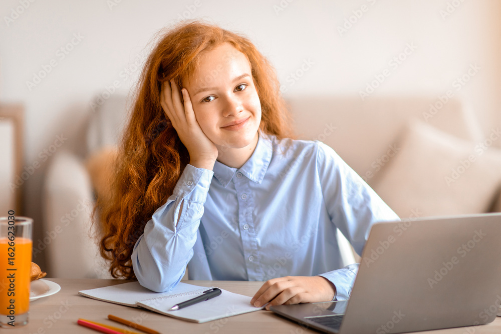 Distance Education. Portrait of smiling red-haired girl sitting at table, using laptop computer and looking at camera, doing homework, studying online at home. E-learning And Knowledge Distance Education. Portrait of smiling red-haired girl sitting at table, using laptop computer and looking at camera, doing homework, studying online at home. E-learning And Knowledge