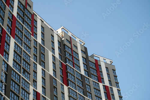 A tall, modern building features a mix of striking red and neutral tones, with numerous large windows reflecting the clear blue sky. The architecture emphasizes a contemporary style