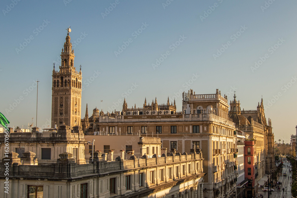 Obraz premium Panoramic elevated view of Seville Cathedral rooftops and historic Giralda bell tower illuminated by warm sunset light in Seville Andalusia Spain.