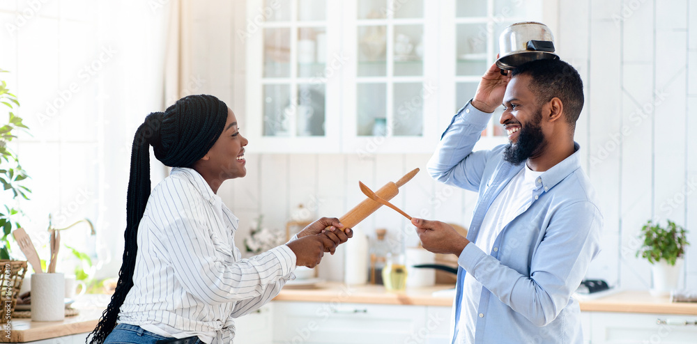 Fototapeta premium Joyful black couple having fun in kitchen, playfully fighting with kitchenware, aiming each other with utensils tools and laughing, young african spouses having enjoying time at home, copy space